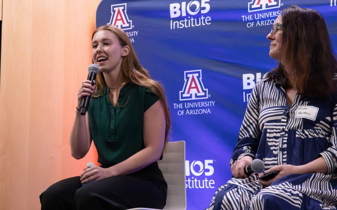 Two individuals seated on stage, speaking at an event at the BIO5 Institute, The University of Arizona. The person on the left holds a microphone and is speaking, while the person on the right listens attentively.