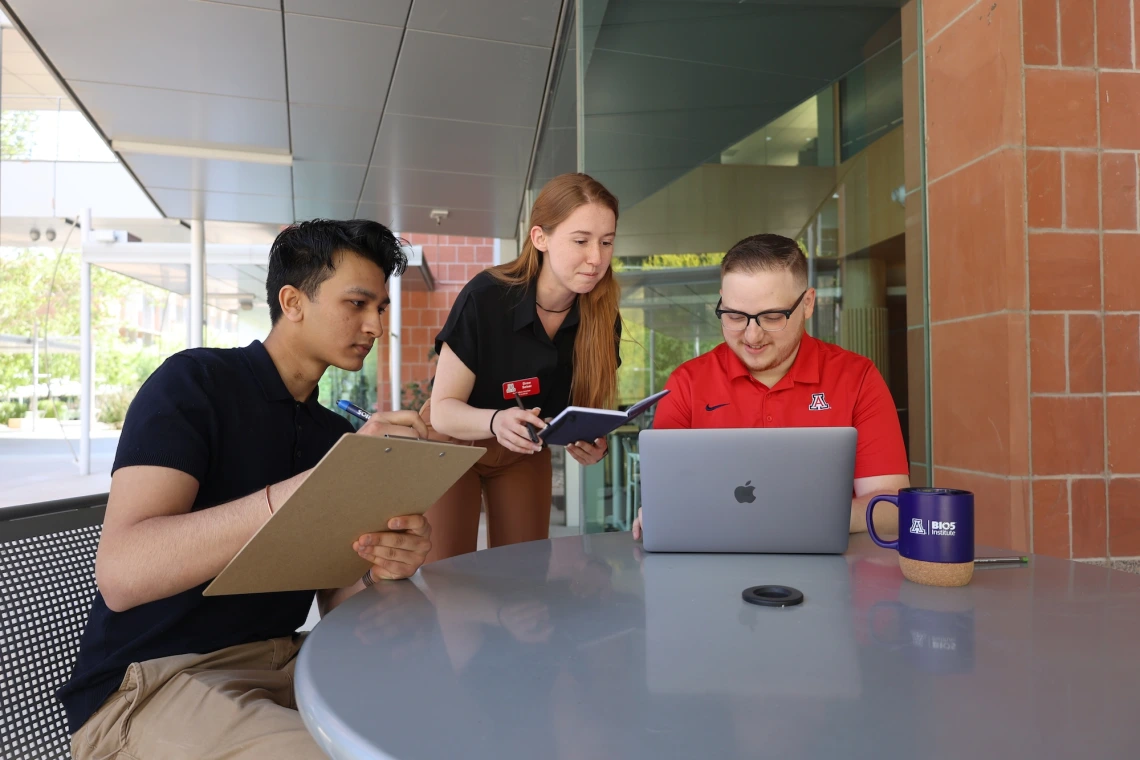 Three individuals in casual business attire work together with digital devices at an outdoor table, with a brick building in the background. One person holds a clipboard, another a tablet, and the third uses a laptop with an Apple logo.