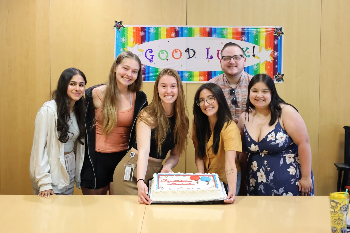 Group of six people smiling in front of a "Good Luck!" banner, with a cake on the table in front of them.