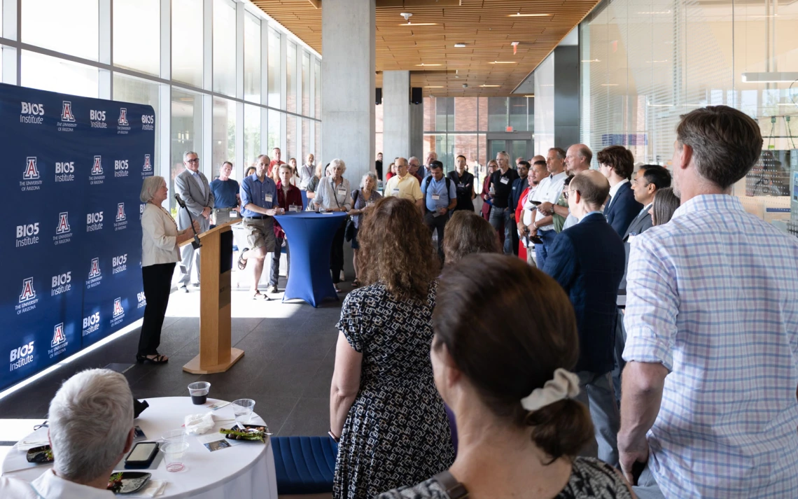A group of people attentively listening to a speaker at a BIO5 event inside a modern building with large windows and a banner displaying the BIO5 logo.