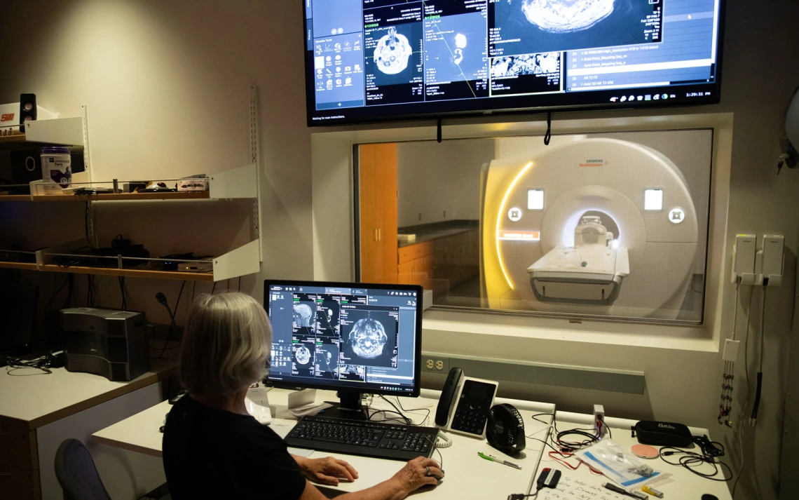 A medical professional is working at a computer monitoring station with multiple screens displaying brain scans, in front of an MRI machine.