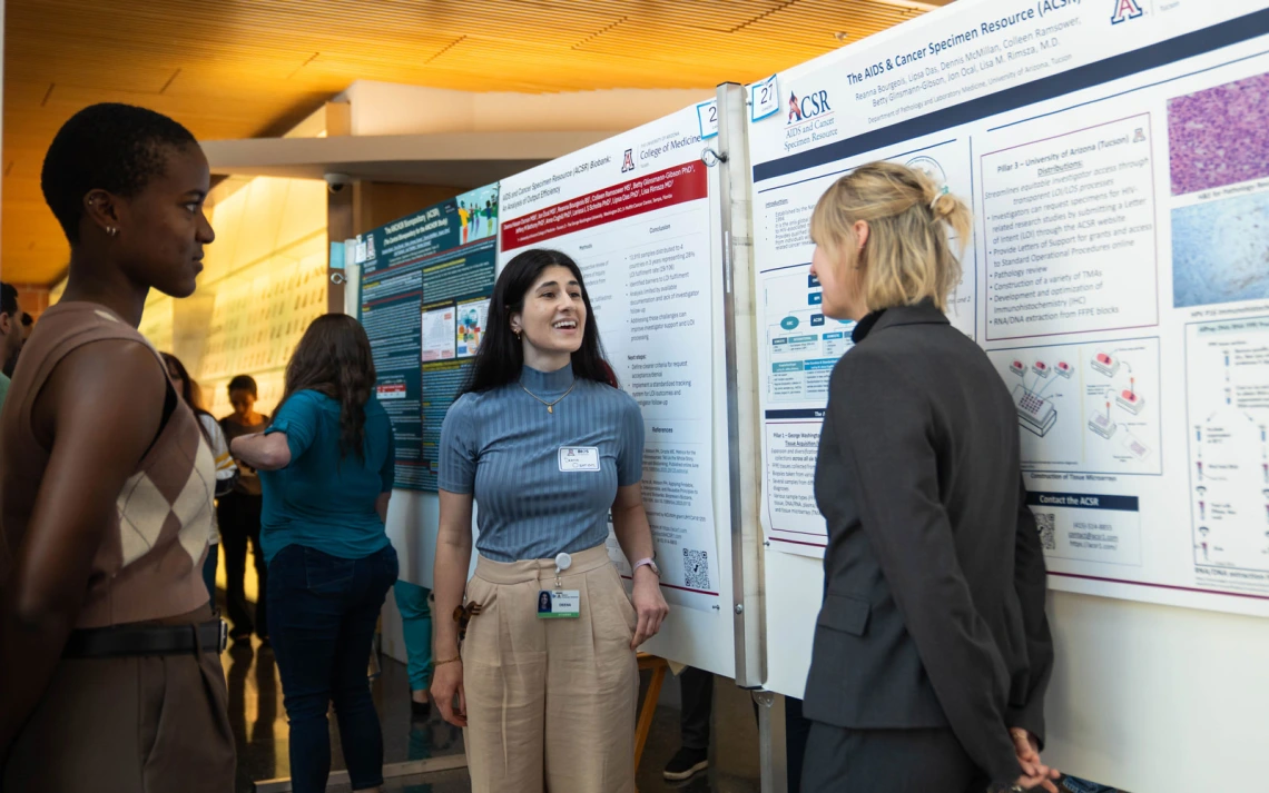 Two individuals discussing research posters at an academic conference, one smiling and engaging with the other who is attentively listening. The backdrop features detailed scientific posters with charts and text.