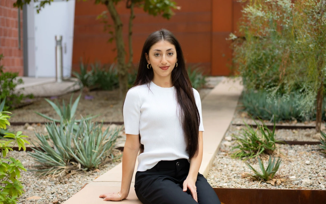 Person sitting on a concrete bench in a garden with various plants and a modern building in the background.