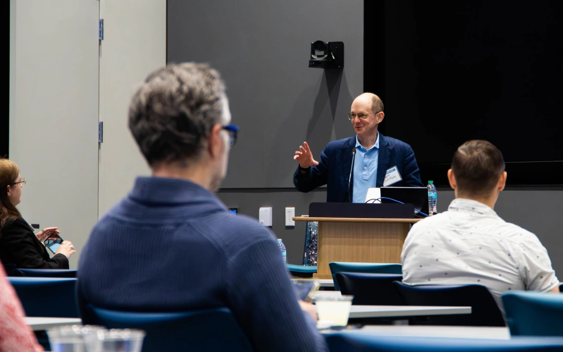 A person presenting to a small group in a modern conference room. The presenter is standing near a digital screen, interacting with the seated audience.