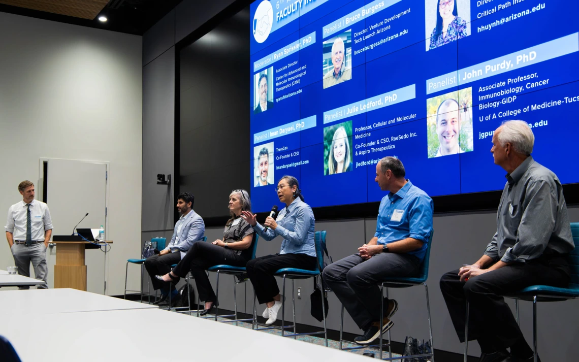 Panel discussion at a conference with six participants seated in front of a digital screen displaying their names and titles. One panelist is speaking while the others listen.