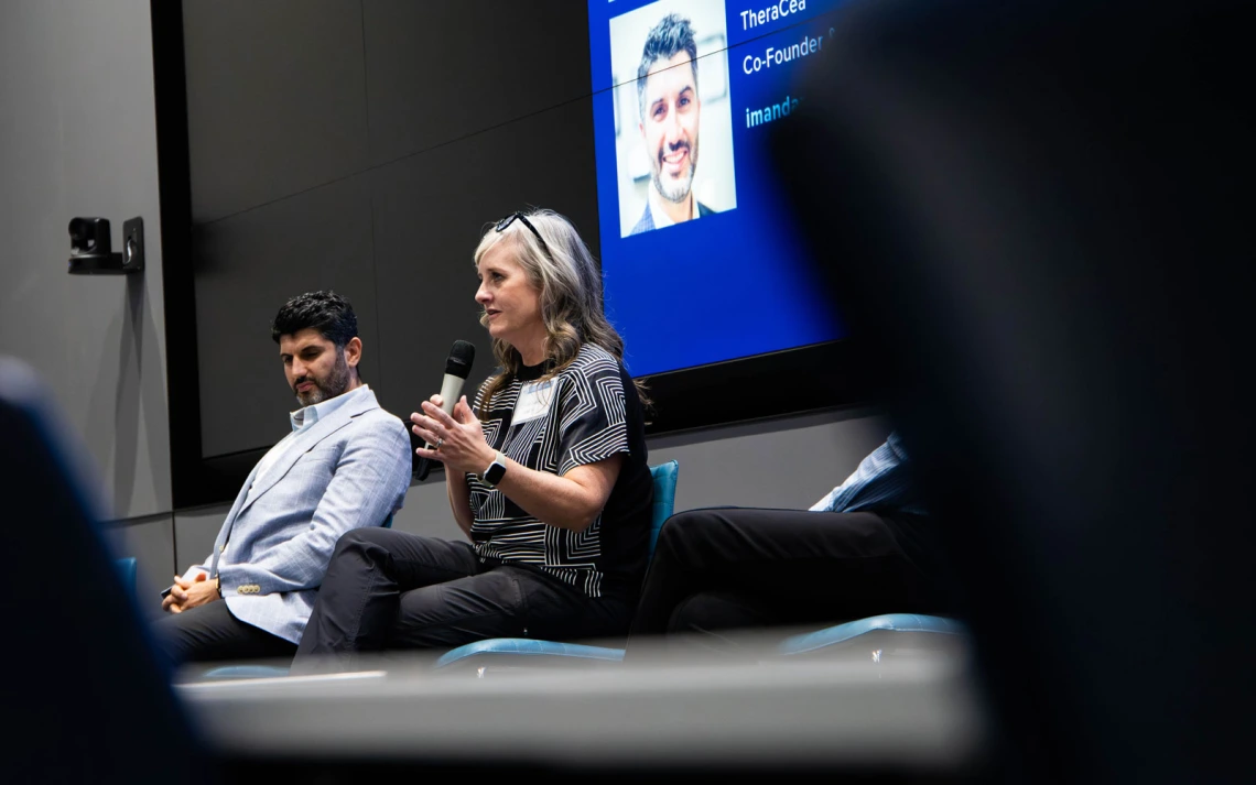 Two people seated in a conference room; one is speaking into a microphone. A presentation screen in the background displays another person's image.