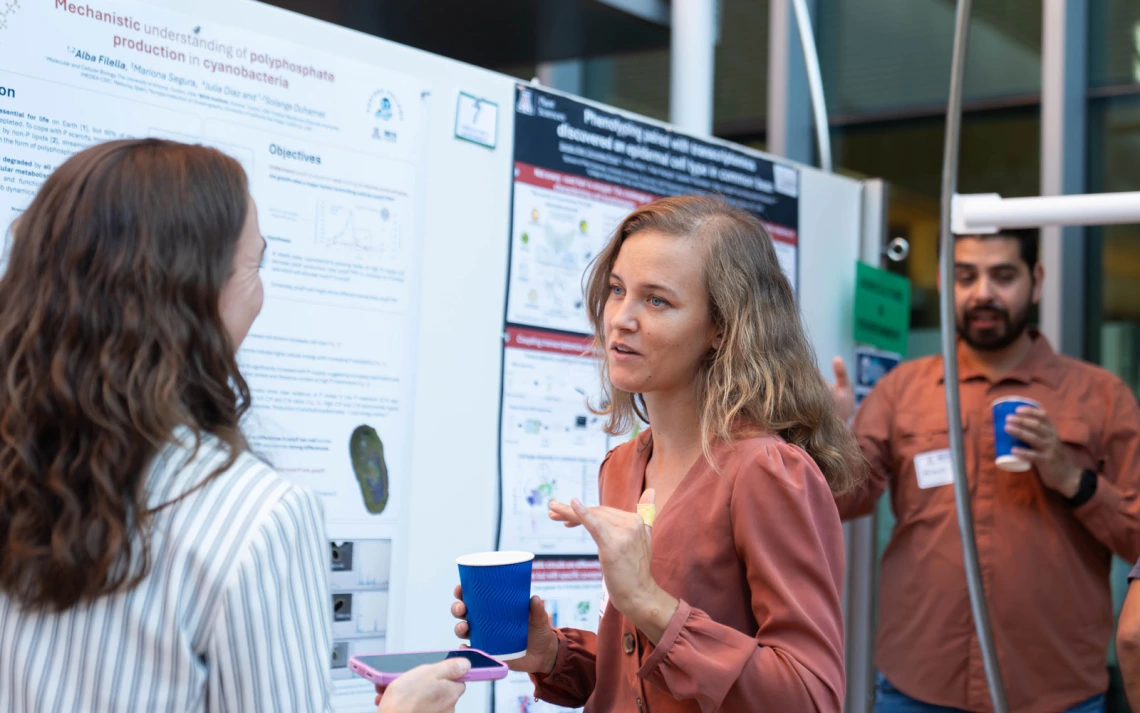 Individual presenting research a poster at a scientific conference, while engaging in conversation with a listener. Another attendee observes in the background.