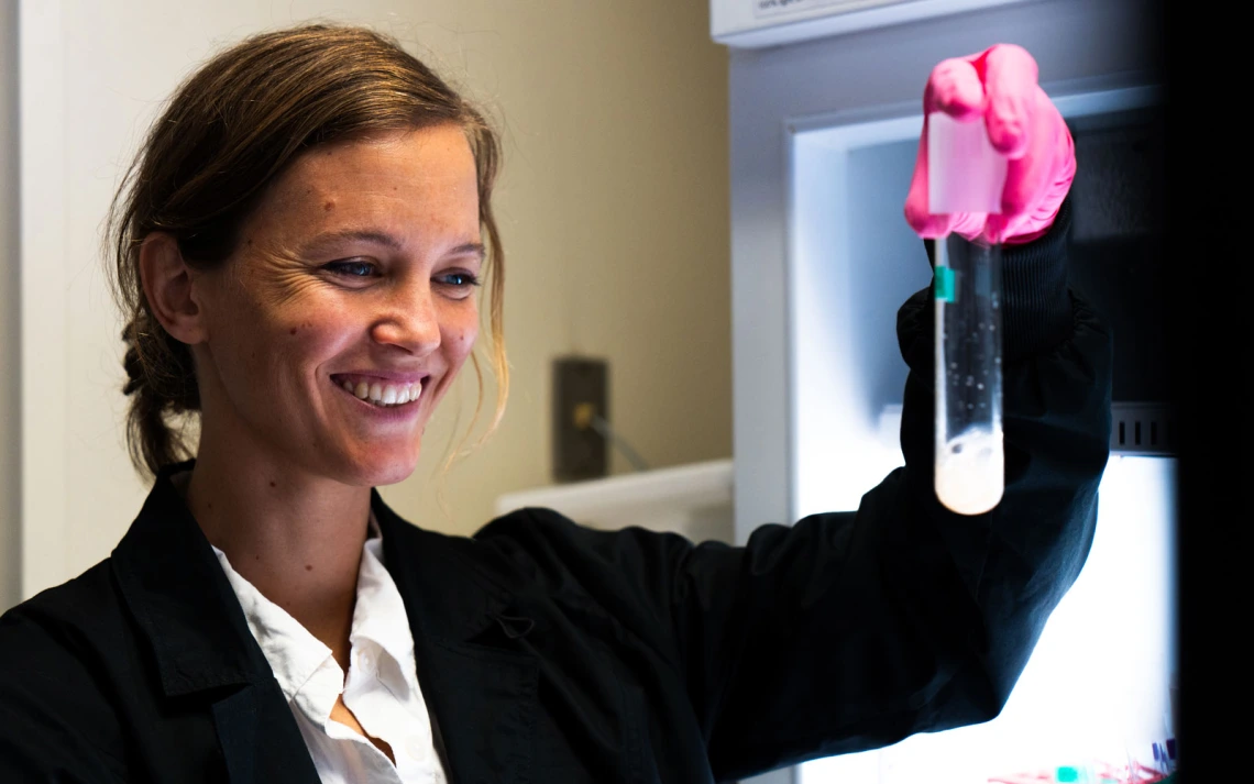 A scientist in a lab coat examines a test tube with a pink substance under a light, smiling with satisfaction.