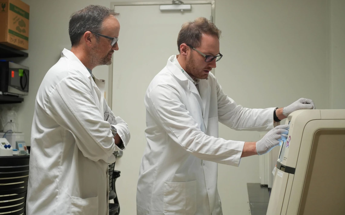 Two individuals in lab coats are in a laboratory setting, with one person demonstrates a piece of scientific equipment while the other observes.