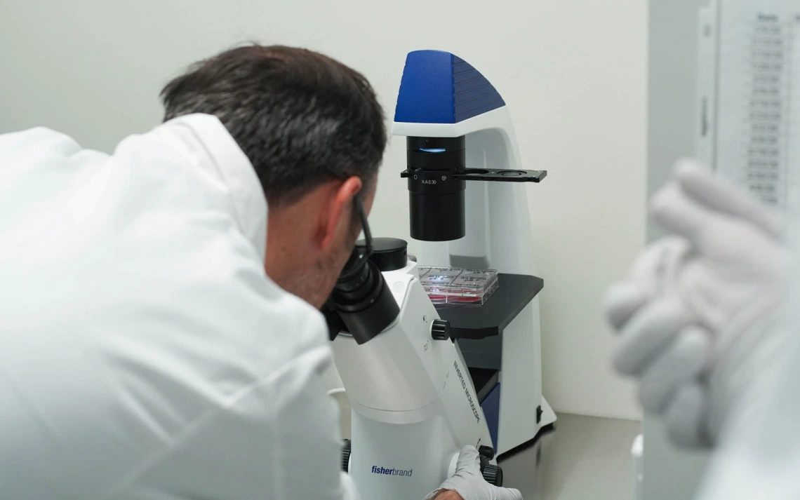 A person in a lab coat uses a microscope to examine a specimen in a lab setting.
