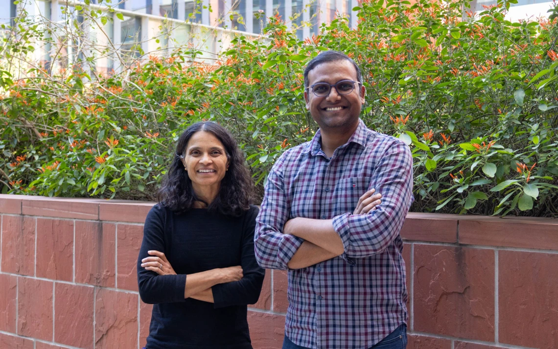 Two people standing in front of a brick planter with green foliage and small orange flowers. 
