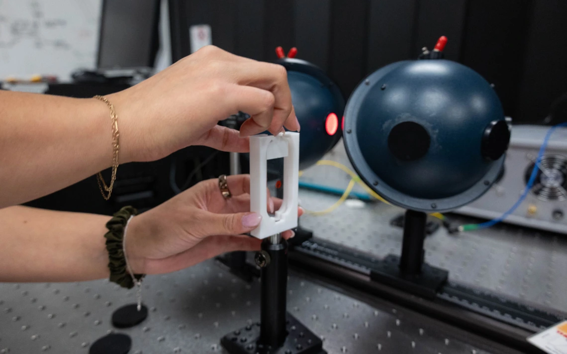 A person holding a small device in front of a setup involving two blue spherical instruments with lenses, likely part of an optical experiment, on a laboratory table.
