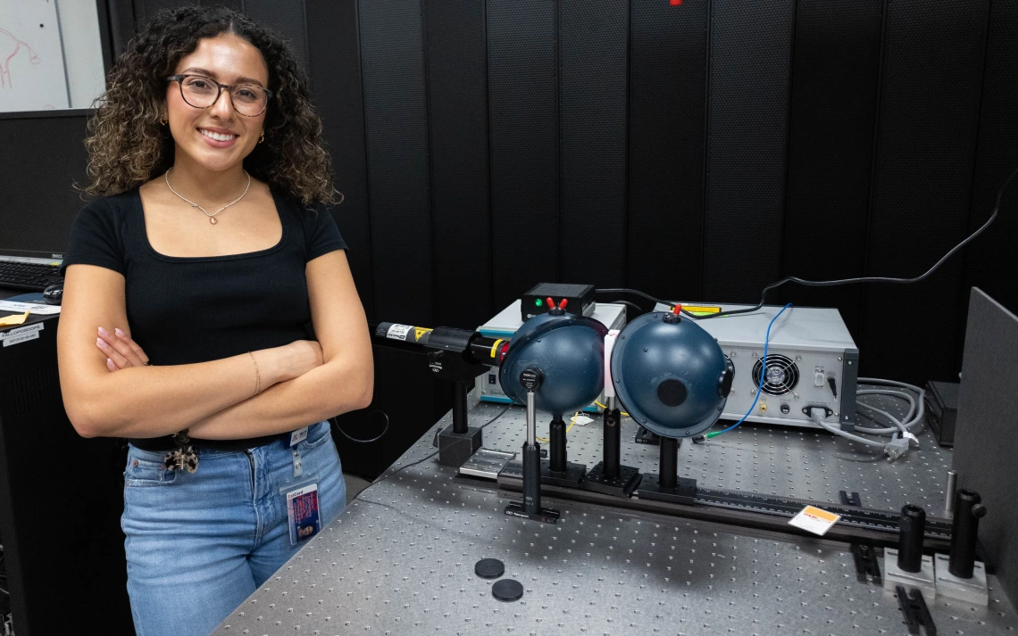 A person with curly hair and glasses stands smiling next to a laboratory setup involving lasers and optical equipment. 