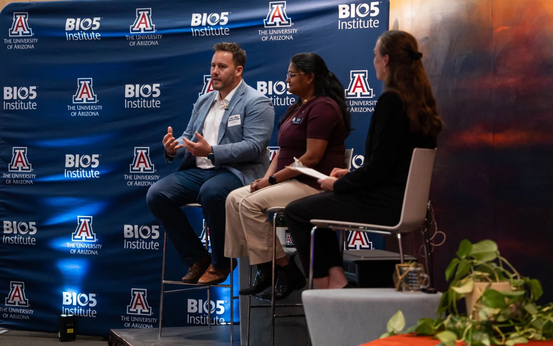 Three people are seated on stools during a panel discussion at the BIO5 Institute. The backdrop features logos for BIO5 Institute and the University of Arizona. One person is speaking, while the others listen attentively. A table with plants is in the foreground.