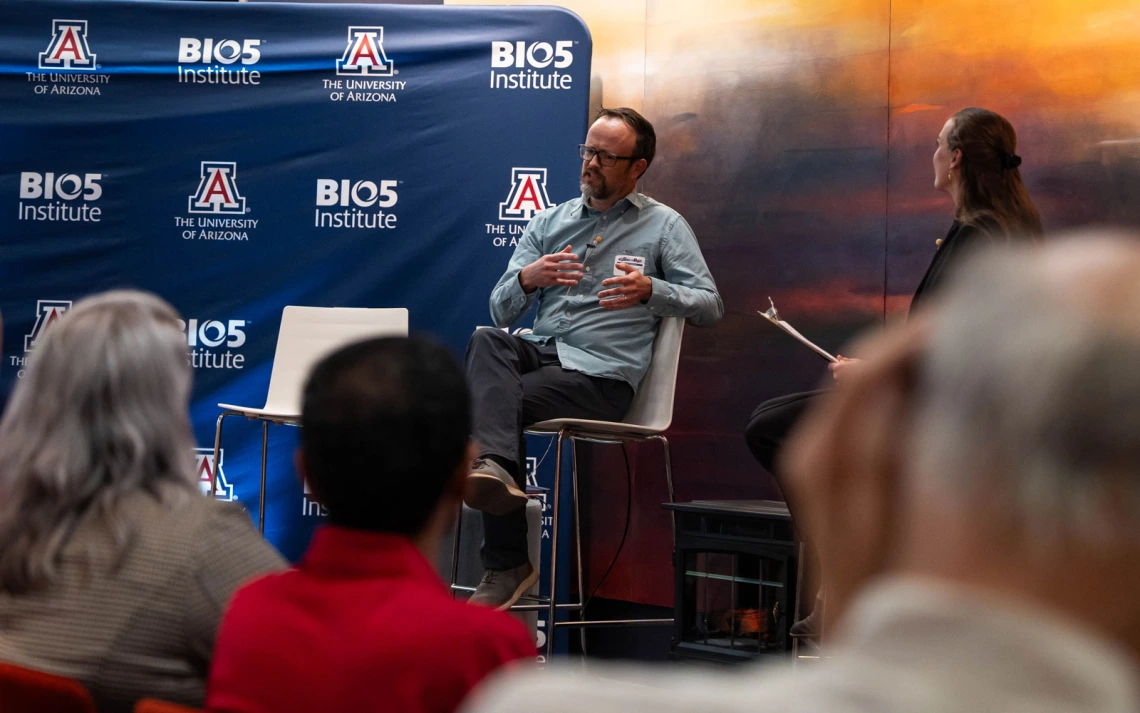 A person sits on a chair, speaking on a stage with a backdrop displaying "The University of Arizona" and "BIO5 Institute" logos. Another individual with a clipboard is seated nearby, facing the speaker. Audience members are visible in the foreground.