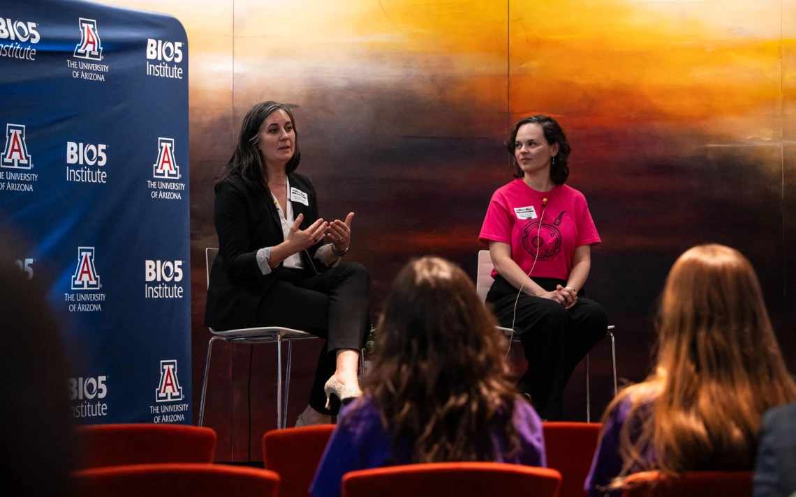 Two individuals are seated on stage at an event. One is speaking while the other listens. Behind them, a backdrop displays logos for "BIO5 Institute" and "The University of Arizona." The audience is visible in the foreground.