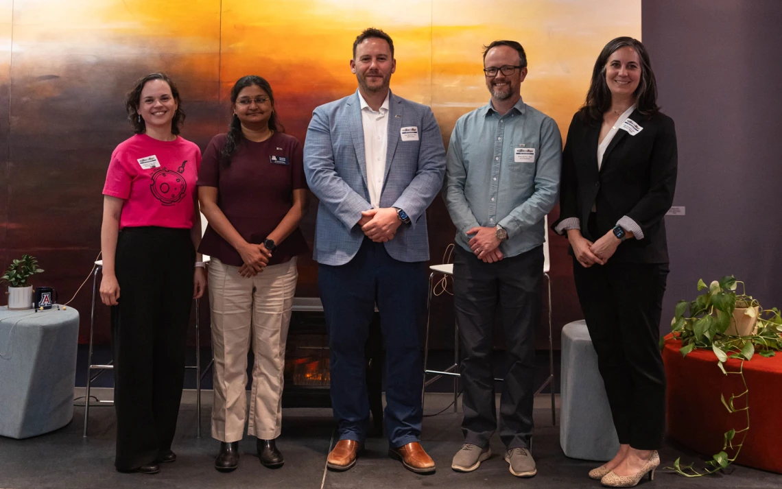 Five people stand in a row, smiling, in front of a large abstract mural, dressed in business casual attire and each has a name tag.