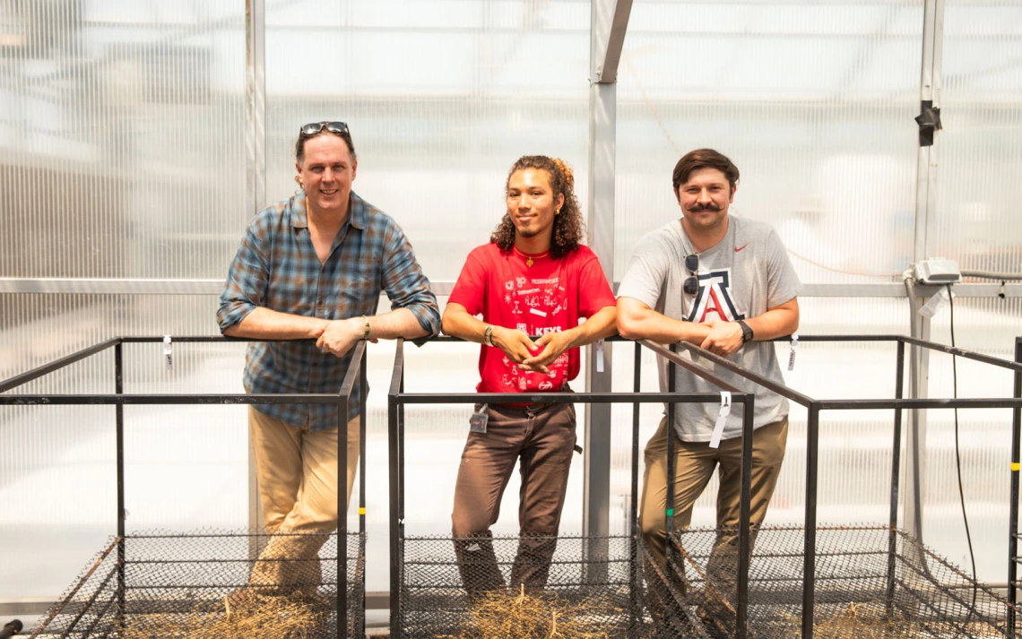 Three individuals stand inside a greenhouse, leaning on metal railings. 
