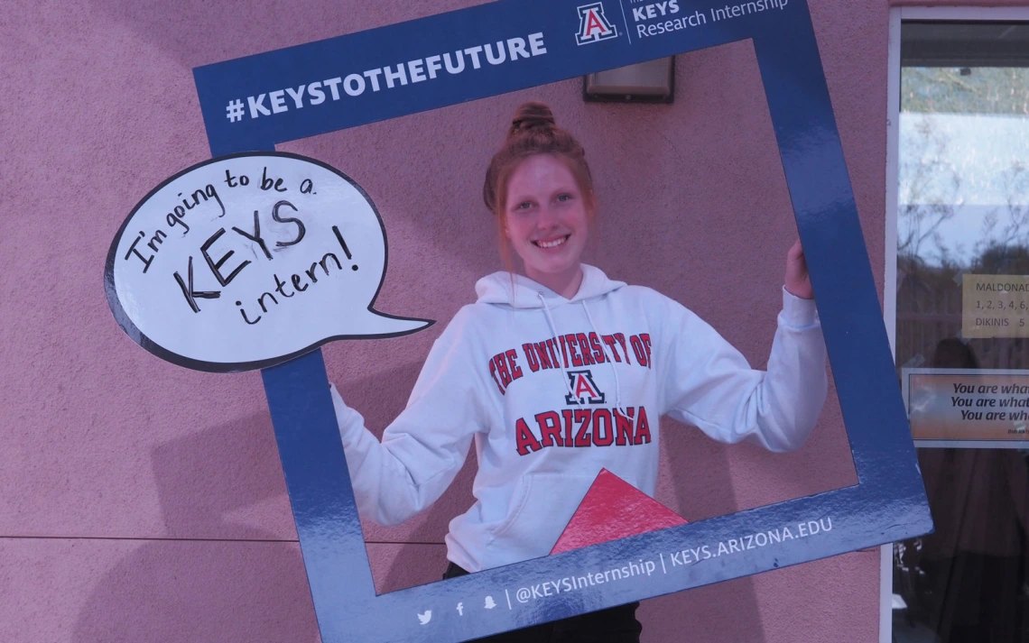 A person smiling and holding a dark blue frame that reads "#KEYSTOTHEFUTURE" and "University of Arizona KEYS Research Internship."