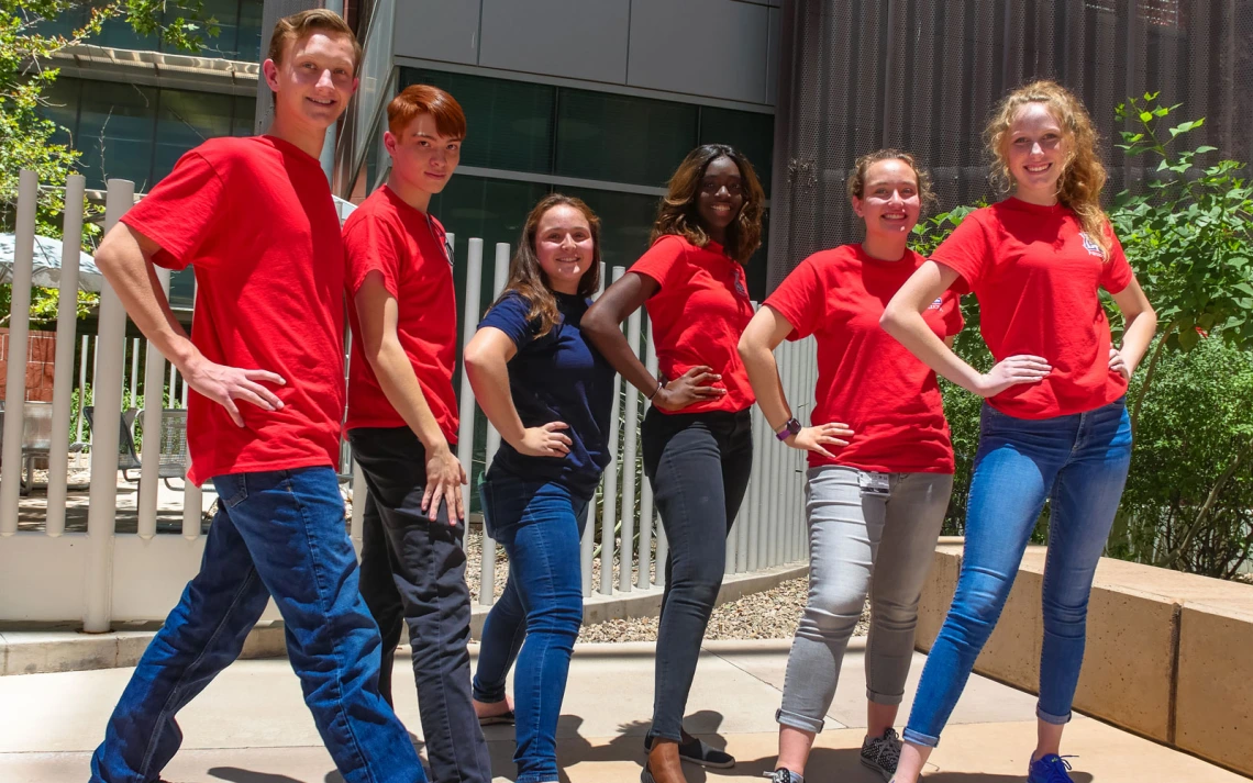 A group of six people standing in a line outdoors, all striking similar poses with hands on hips. 