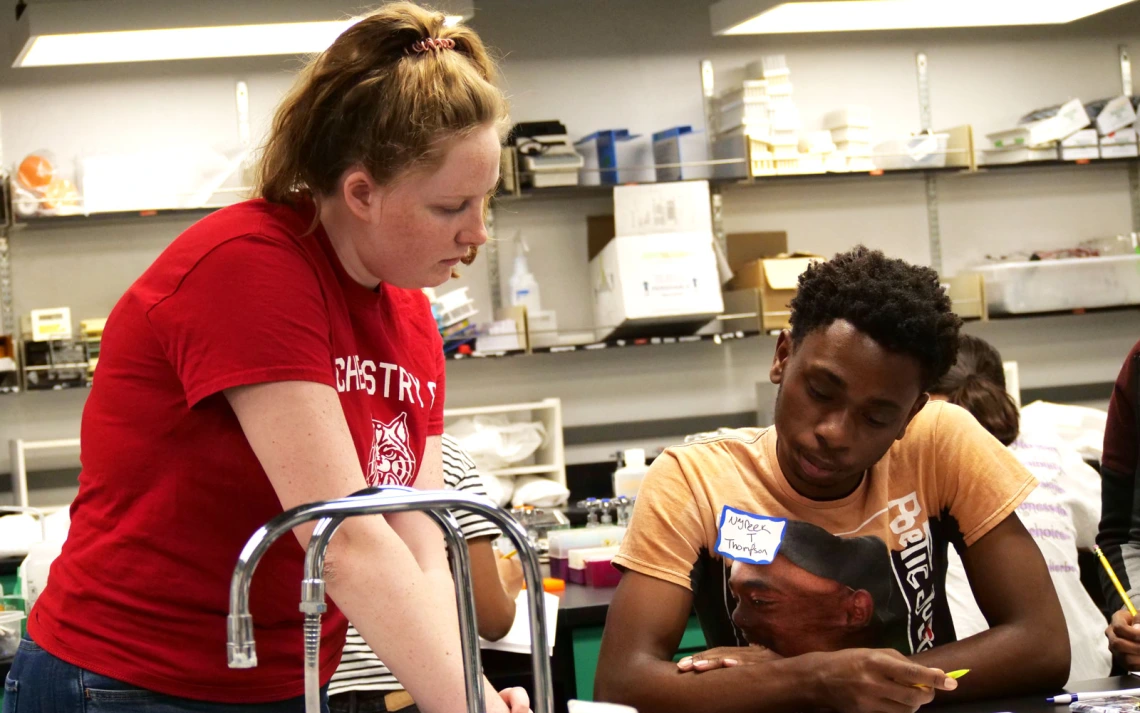 A person in a red shirt is assisting another person sitting at a table in a lab setting. 
