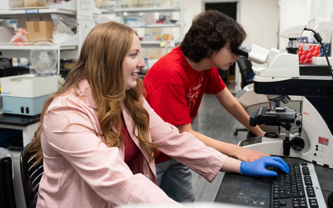 Two people are in a laboratory. One is looking into a microscope while the other is using a computer keyboard.