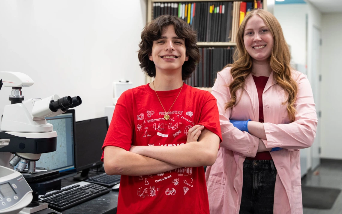 Two people stand smiling in a laboratory setting. One is wearing a red shirt with graphics, and the other is in a light coat with blue gloves.