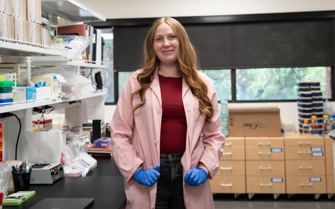 A person stands confidently in a laboratory setting, wearing a pink lab coat and blue gloves. The laboratory is filled with equipment, shelves, and supplies.