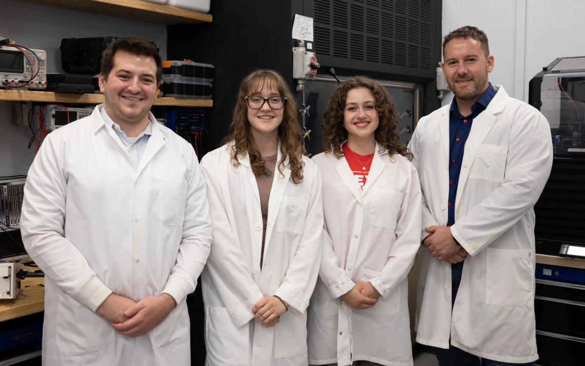 A group of four people in lab coats standing and smiling in a laboratory setting with shelves and equipment in the background.