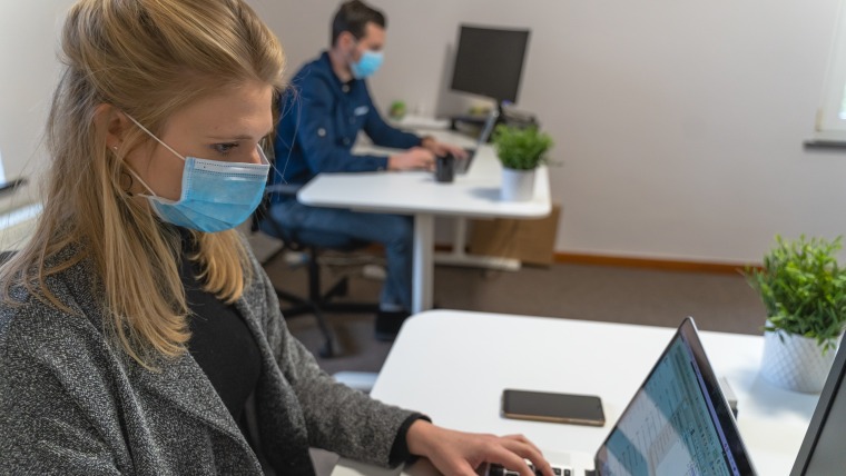 Man and woman using laptops, sitting at separate desks wearing blue surgical masks