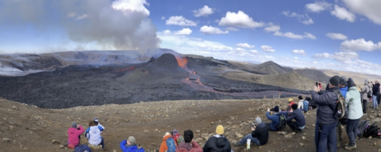 A volcano erupts in the background as researchers and onlookers watch in the foreground