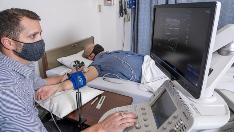 Dr. Dallin Tavoian uses an ultrasound probe to obtain an image of an artery in the participant's upper arm. 
