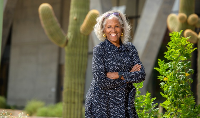 Dr. Juanita merchant wearing a dark colored pattered dress, standing outside in front of a cactus