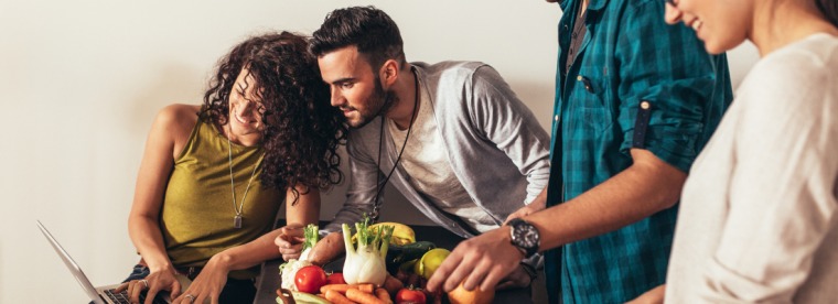 Man and woman looking at a laptop and smiling behind two people handling vegetables