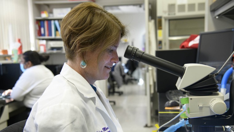 Profile point of view of Helena Morrison wearing a white labcoat in front of a microscope in the lab