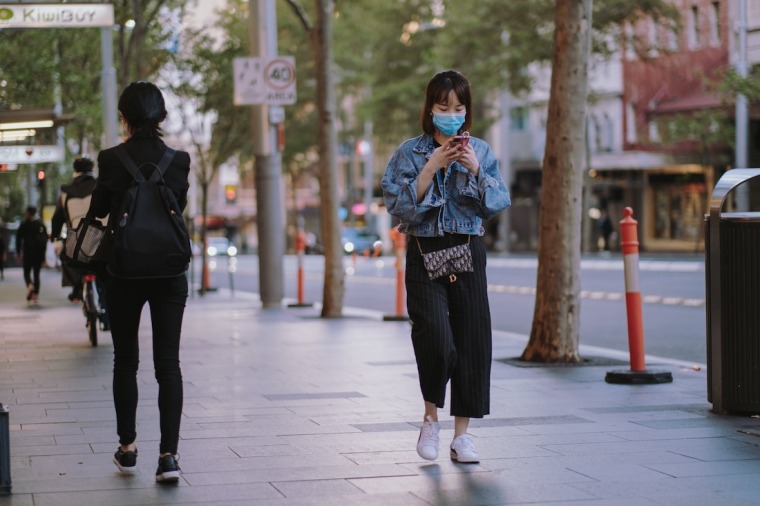 Woman wearing mask and walking while looking at phone.