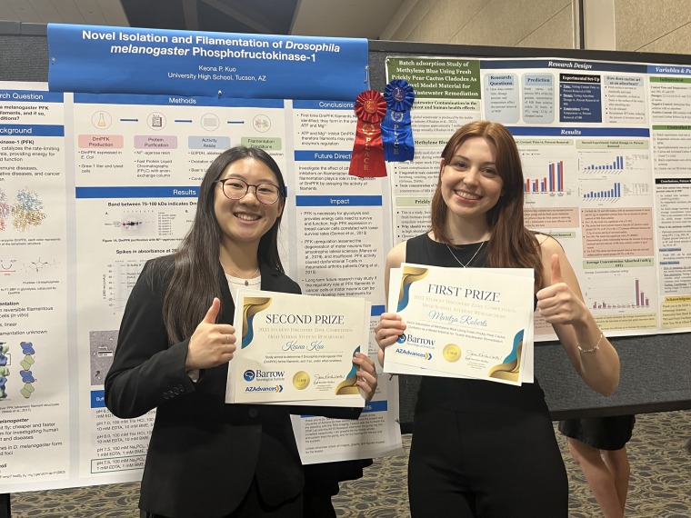 Two young women stand in front of scientific posters and hold their winners' certificates