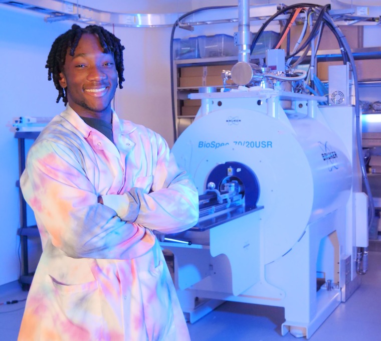 Young black man wearing a tie dye lab coat crosses his arms in front of him. In the background is an MRI machine.