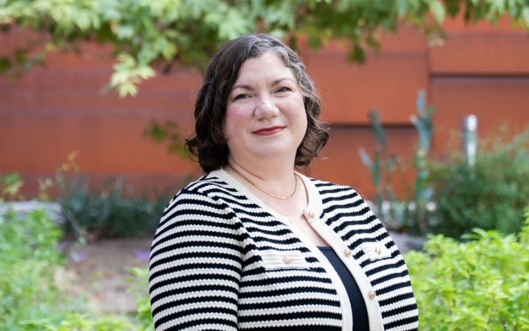 Person smiling and dressed in a striped jacket, standing outdoors with greenery and a red brick wall in the background.