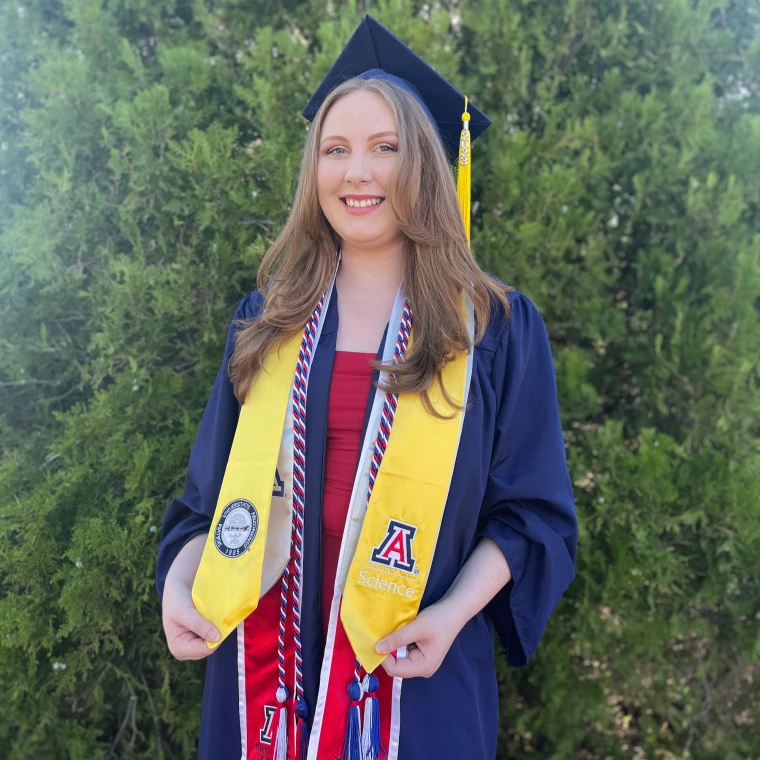 A person wearing a graduation cap and gown is standing in front of green foliage.