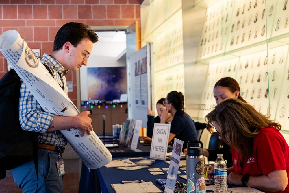 Person holding a poster interacts with another person seated at a booth labeled "Sign in Here" at a professional event.