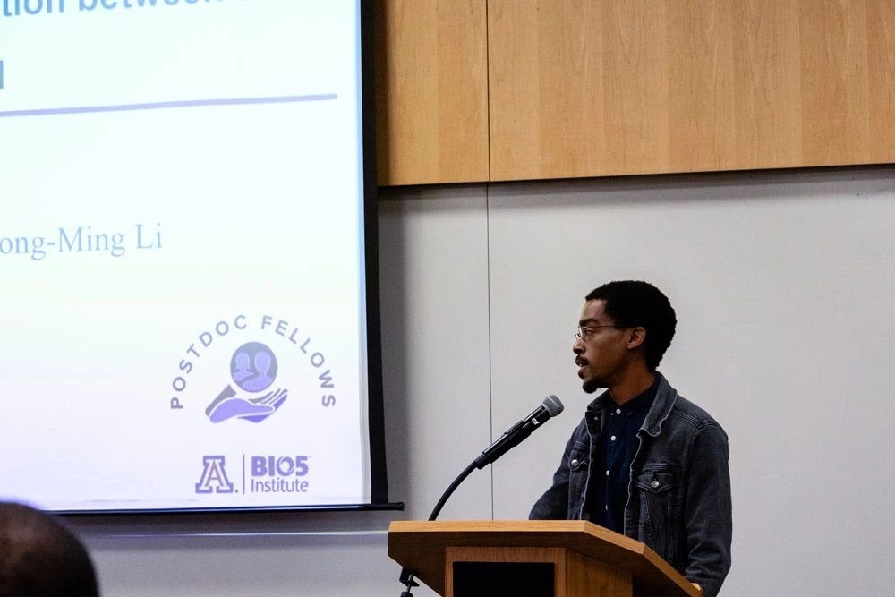 A person presenting at a lecture, standing next to a projector screen with text and the logo of the Postdoc Fellows at the BIO5 Institute