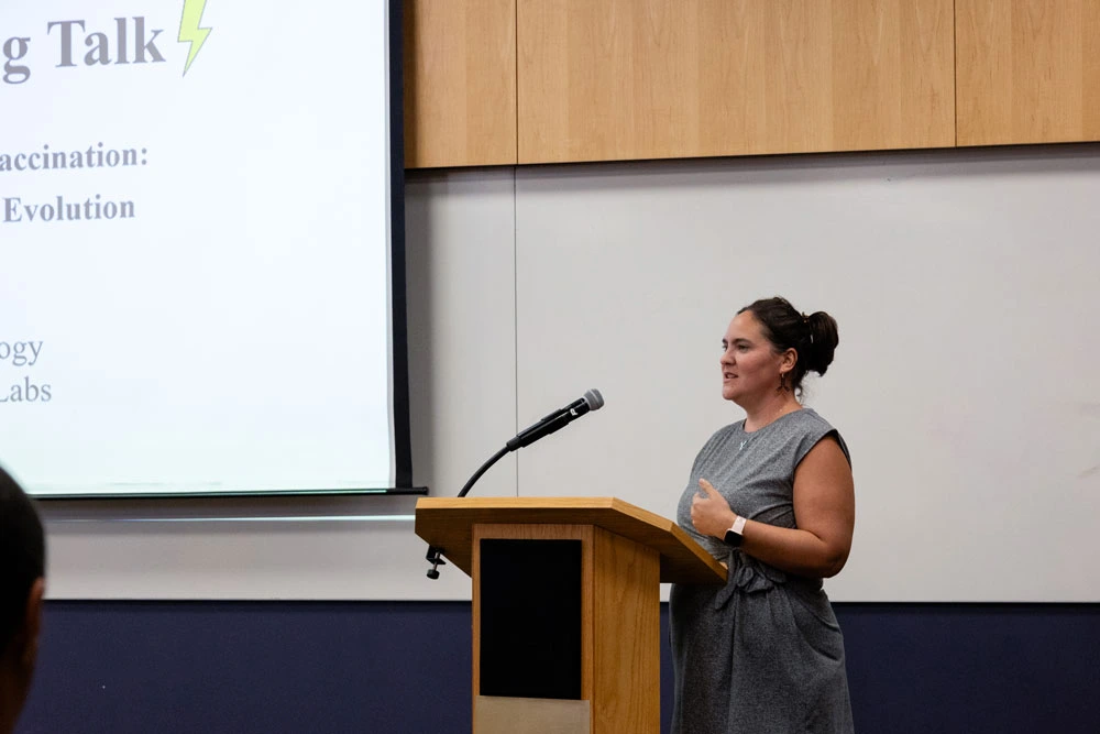 A person presenting at a lectern with a slideshow projected on a screen behind them, in a conference room setting.