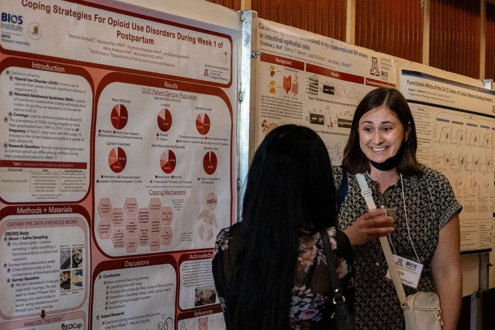 Two individuals at a scientific conference discuss in front of posters, which includes various graphs and text sections like Introduction, Results, and Methods & Materials.