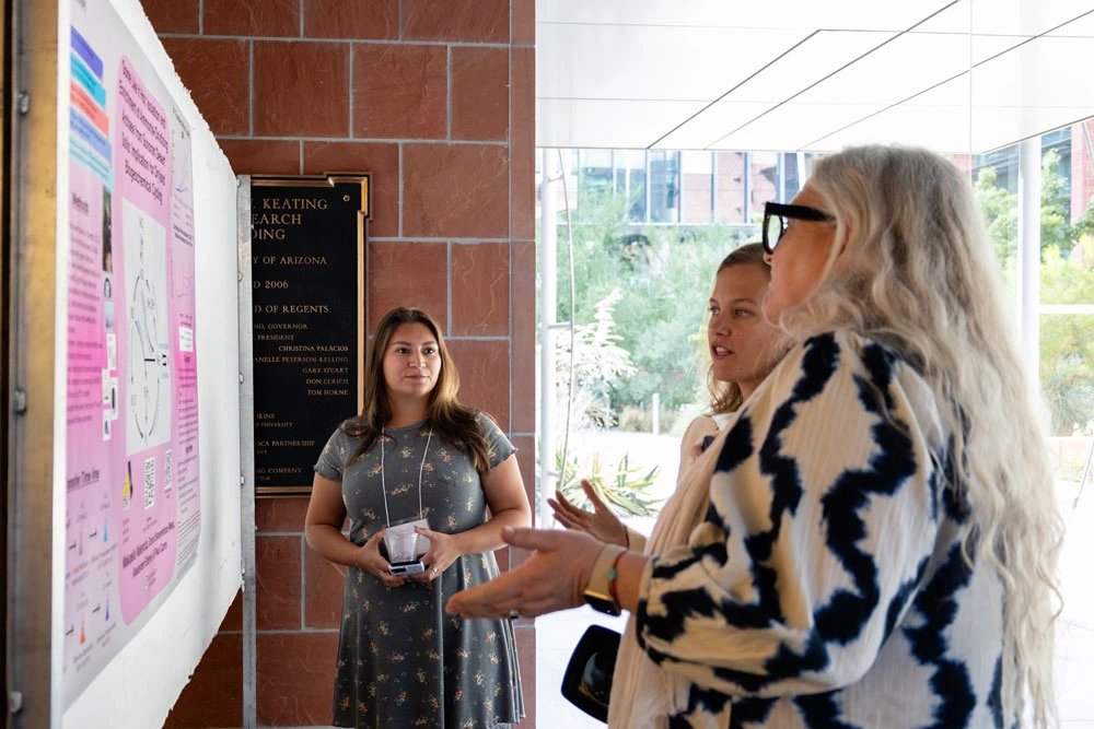 Three people discussing a presentation next to a poster board in a university building. One person is gesturing towards the poster while the other two listen attentively.