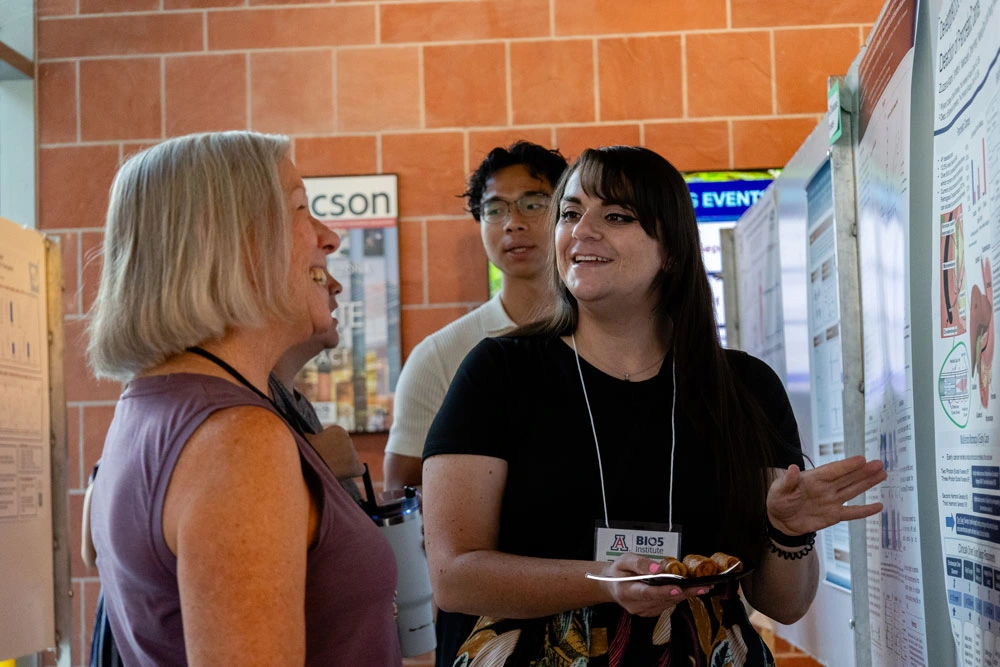 Two individuals engaging in a conversation at a poster presentation session in a brick-walled, indoor setting. One person is gesturing while the other is listening attentively. All are wearing conference badges.