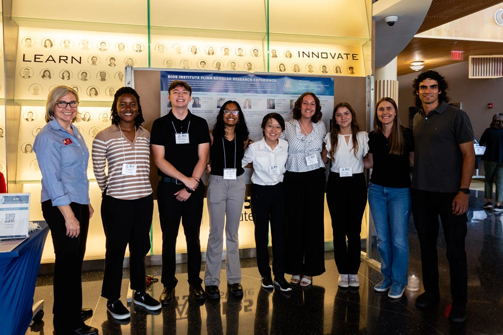 Group of eight individuals standing in front of a display with headshots, smiling and posing for a photo.