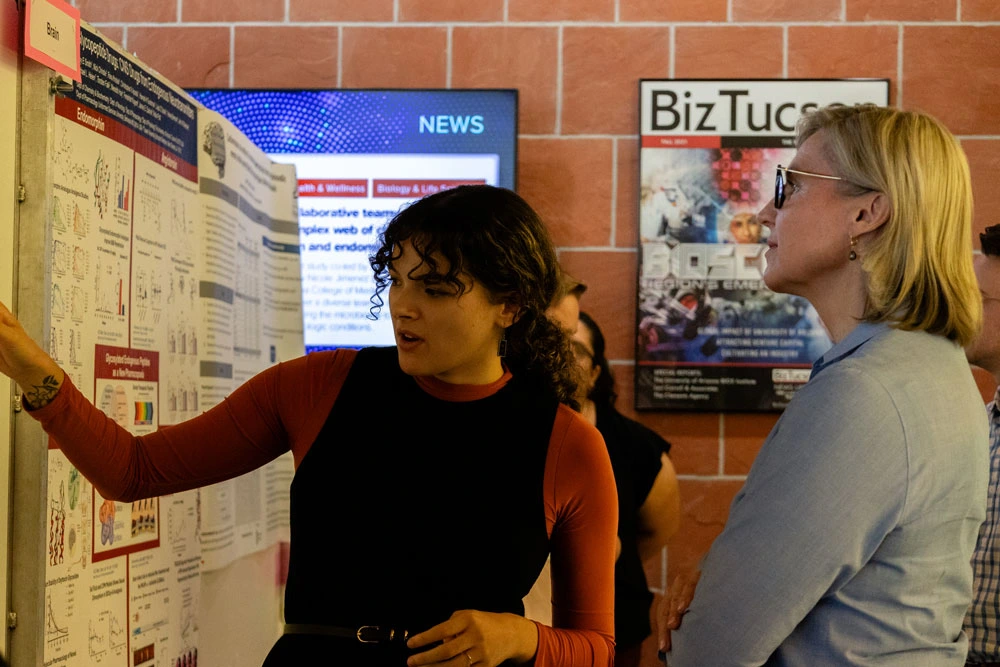 Two individuals examining and discussing a research poster at a conference. One person is pointing at the poster, which shows various charts and text, while the other listens attentively. 