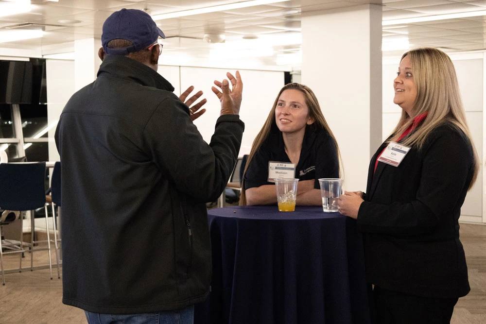 Three individuals engaging in conversation at a networking event, with two listeners wearing name tags and standing behind a table with a drink.