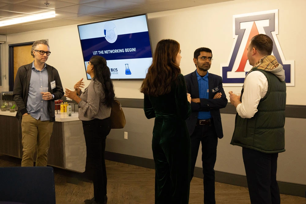 Five individuals are engaged in conversation at a networking event at the University of Arizona, as indicated by the logo on the screen displaying "Let the Networking Begin." 